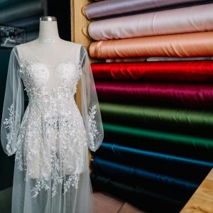 Intricately embroidered white wedding dress on a mannequin surrounded by colorful fabric bolts indoors.