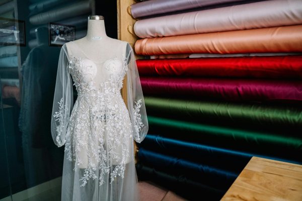 Intricately embroidered white wedding dress on a mannequin surrounded by colorful fabric bolts indoors.